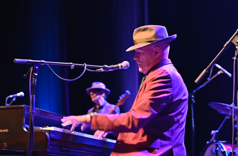 My Leonard Cohen with Stewart D’Arrietta - a side view of a man in a hat with a brim playing a piano on a stage with other musicians.
