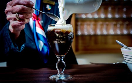 The Irish Coffee Experience, view of a person pouring cream from a jug into a glass with a stem containing a dark coloured drink.