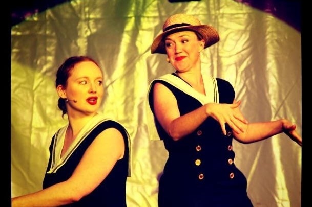 2 women on a stage, on seated at a piano singing, the other dancing beside with gold background.