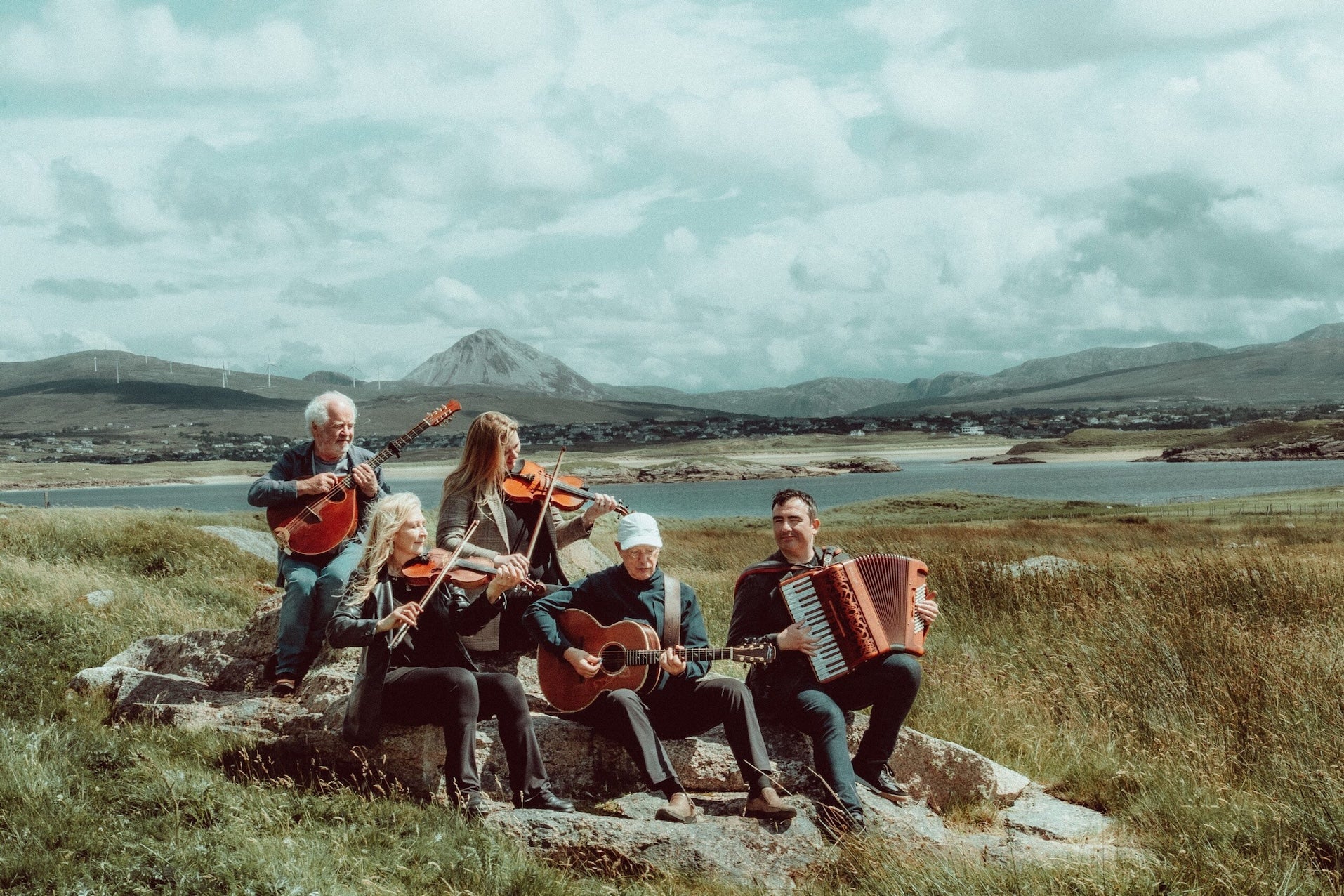 A group of 5 musicians seated on rocks playing with scenic view of mountains and water behind.