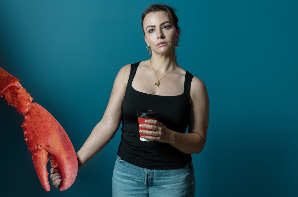 Ross and & Rachel by James Fritz, an unhappy looking woman with a takeout cup of coffee is holding the large claw of a large lobster.