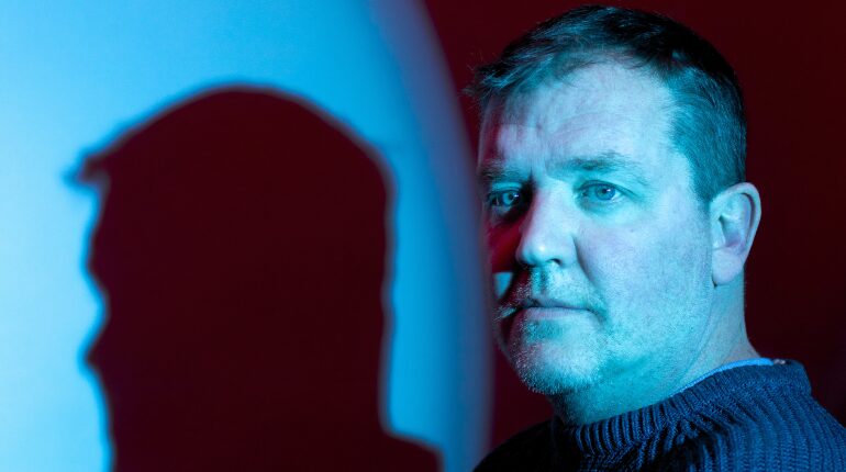 A serious looking man lit in blue with shade of his head cast to one side of him.