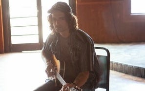 A serious looking man seated on a chair with a guitar with back to the light from a doorway