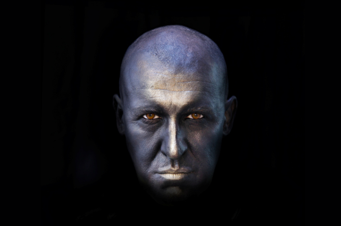 UNDERNEATH written and performed by Pat Kinevane - a bald man's face in silver colour surrounded by blackness.