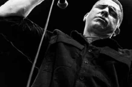 Black and white image looking up at a serious looking man, holding a mic.