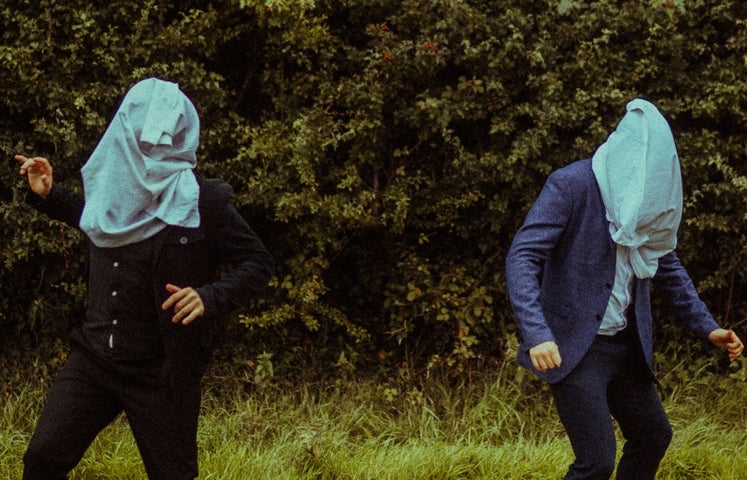 2 people outdoors with hedge in background, standing apart with large, pale blue garments over their heads.