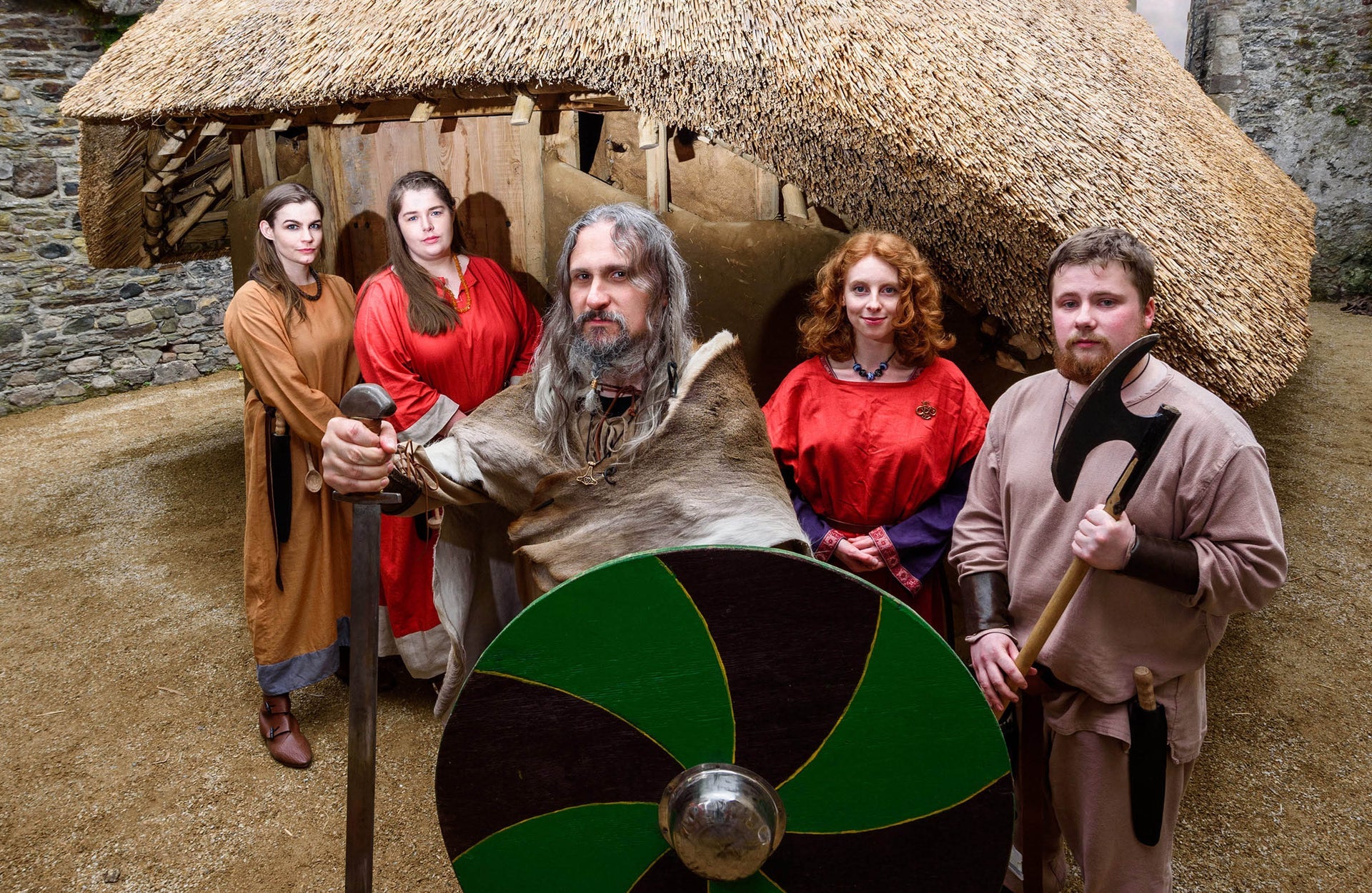 People dressed at Vikings holding weapons and a green and black shield outside a thatched replica Viking house