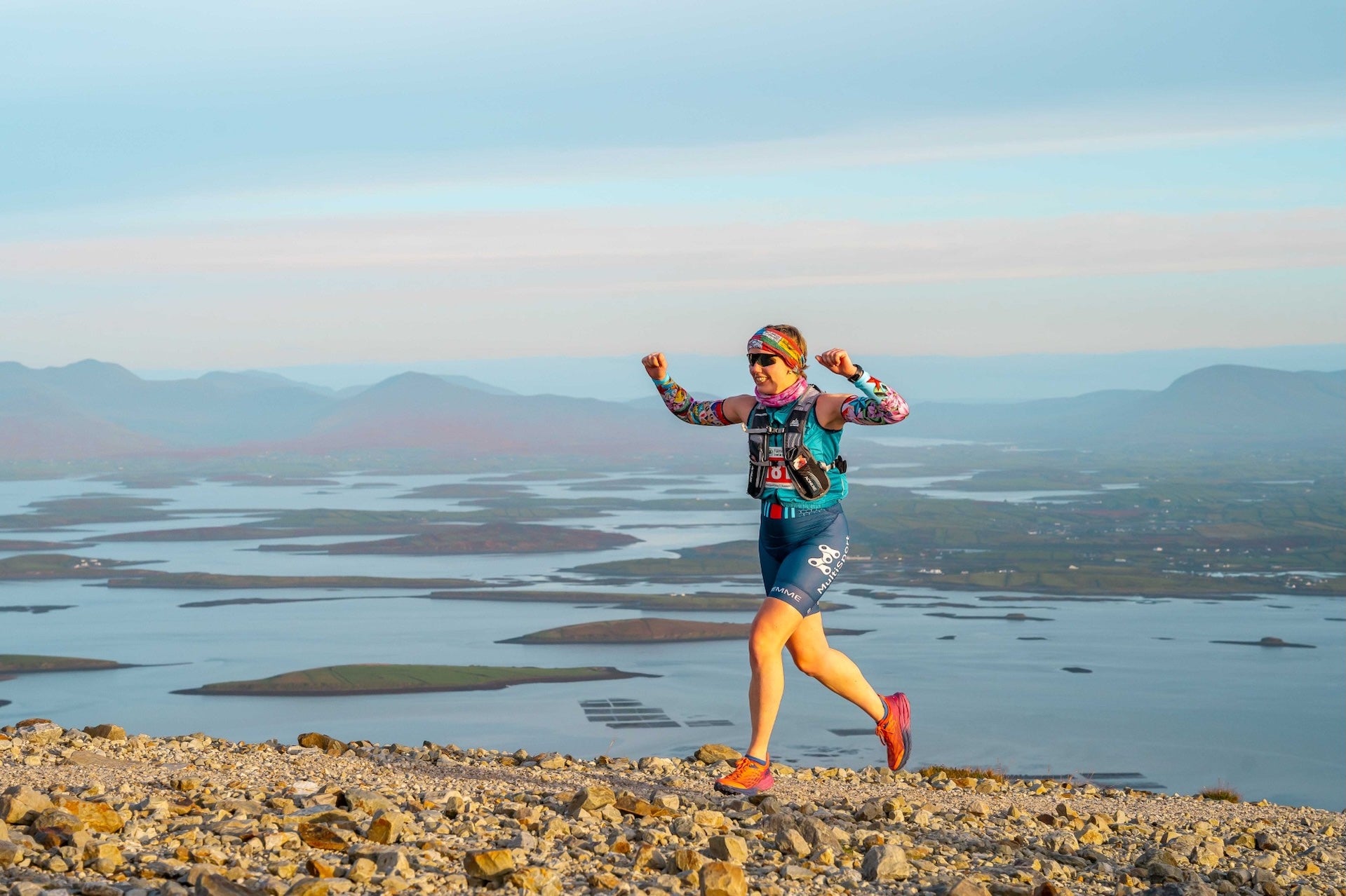 Westport Sea 2 Summit Adventure Race - a person running with arms raised, on top of a high hill with water and hills below in the background.