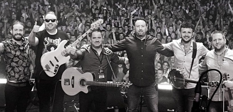 A line of 6 smiling men on a stage with their backs to a large audience.