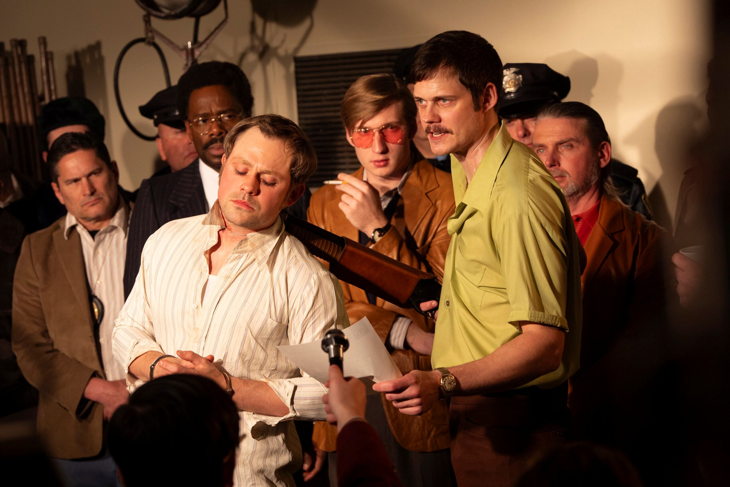 A man is pointing a shotgun at a bound man while holding a sheet of paper. Someone is holding a microphone towards them and there are other men in the background.