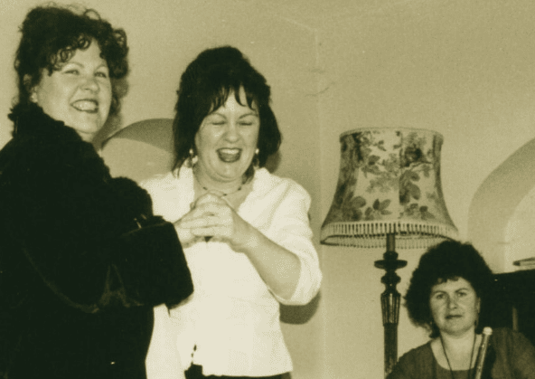 Old photo of 2 women standing and laughing with seated woman holding pipes.