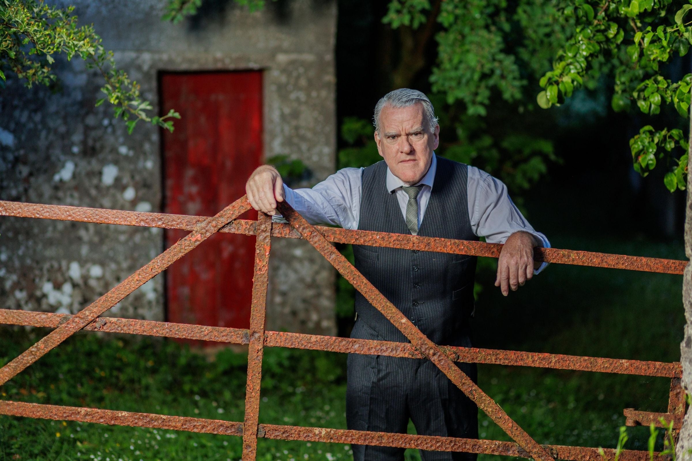 A rather cross looking older man in shirt, tie and waist coat is leaning his arms on a rusty gate