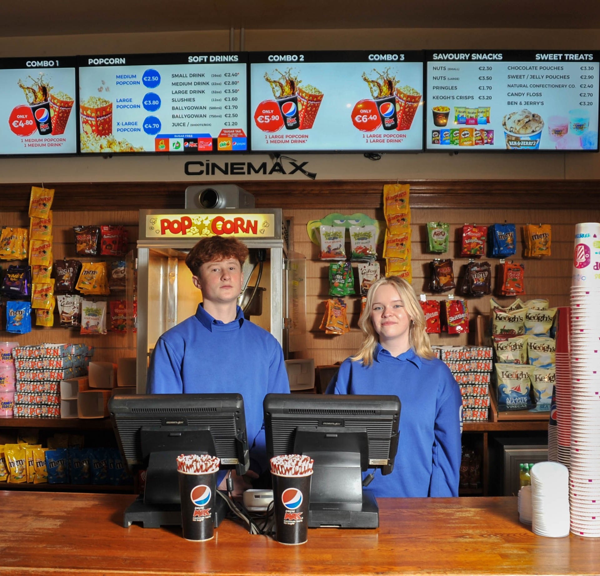 Two people standing behind a wooden counter with packets of sweets a popcorn machine and light up sign advertising cinema snacks