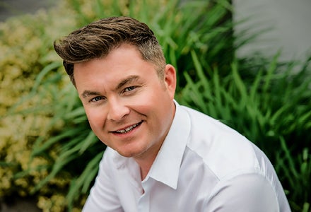 A slightly smiling man in white shirt with grasses in the background.