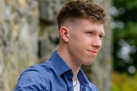 A gently smiling young man with blurry greenery in the background.