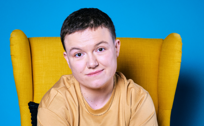 Comedian Sinead Walsh, gently smiling, seated in a yellow, highback armchair.