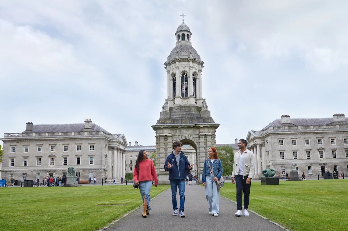 3 people walking on a path with a tour guide talking, impressive grand, stone buildings and tour in the background.