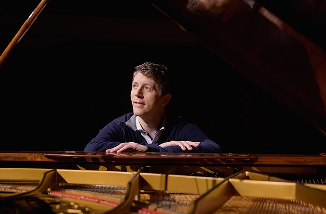 Looking across an open grand piano with a man sat at the keyboard end, looking away, resting his hands on top
