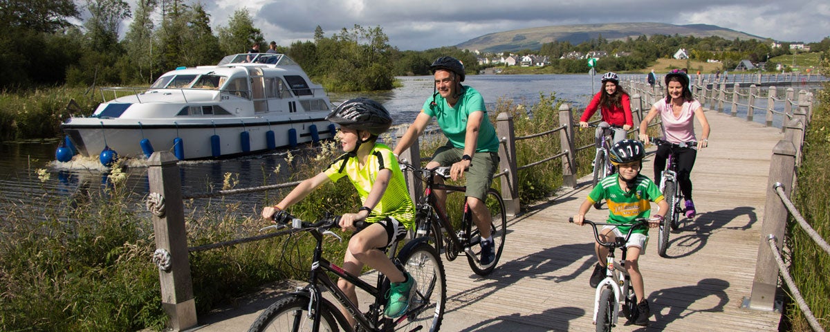 Two adults and three children on bikes on a boardwalk adjacent to a river with a boat on it