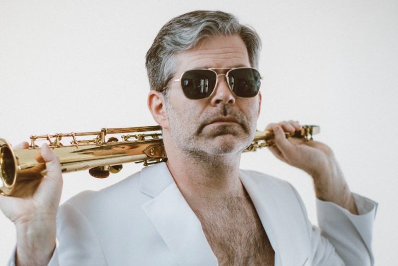 A man in sunglasses and a white open jacket showing a hairy chest is holding a gold coloured wind musical instrument over his shoulders.