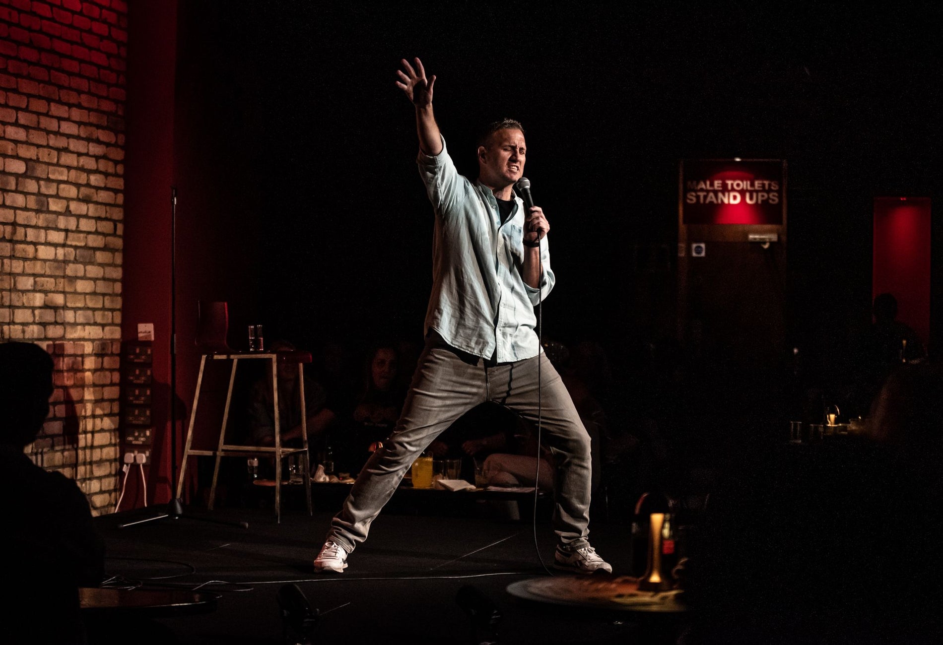 A comedian on stage holding a microphone with his right arm and leg extended