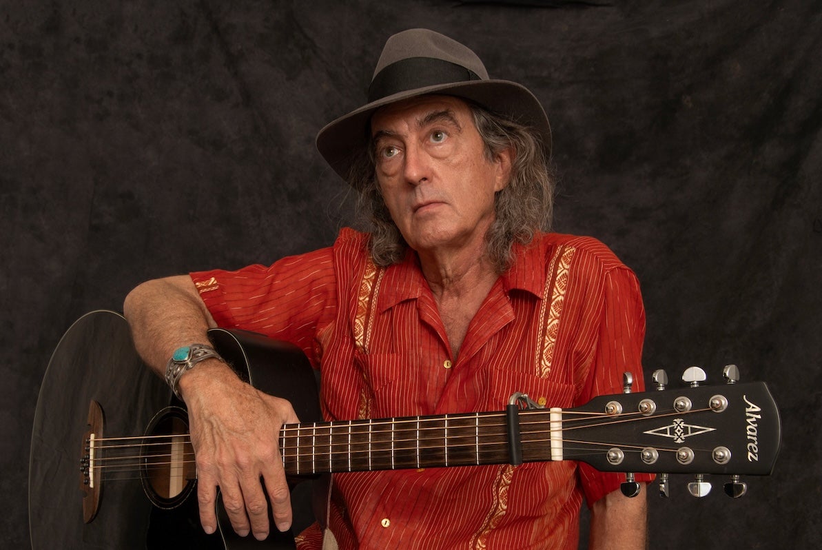 A man in red shirt and grey hat is holding a guitar.