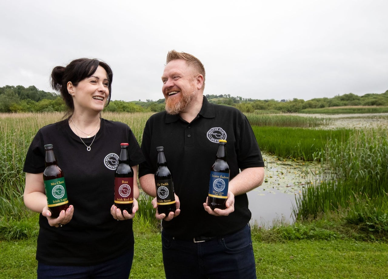 Two people showcasing a selection of beers against the backdrop of a lakeside wetland