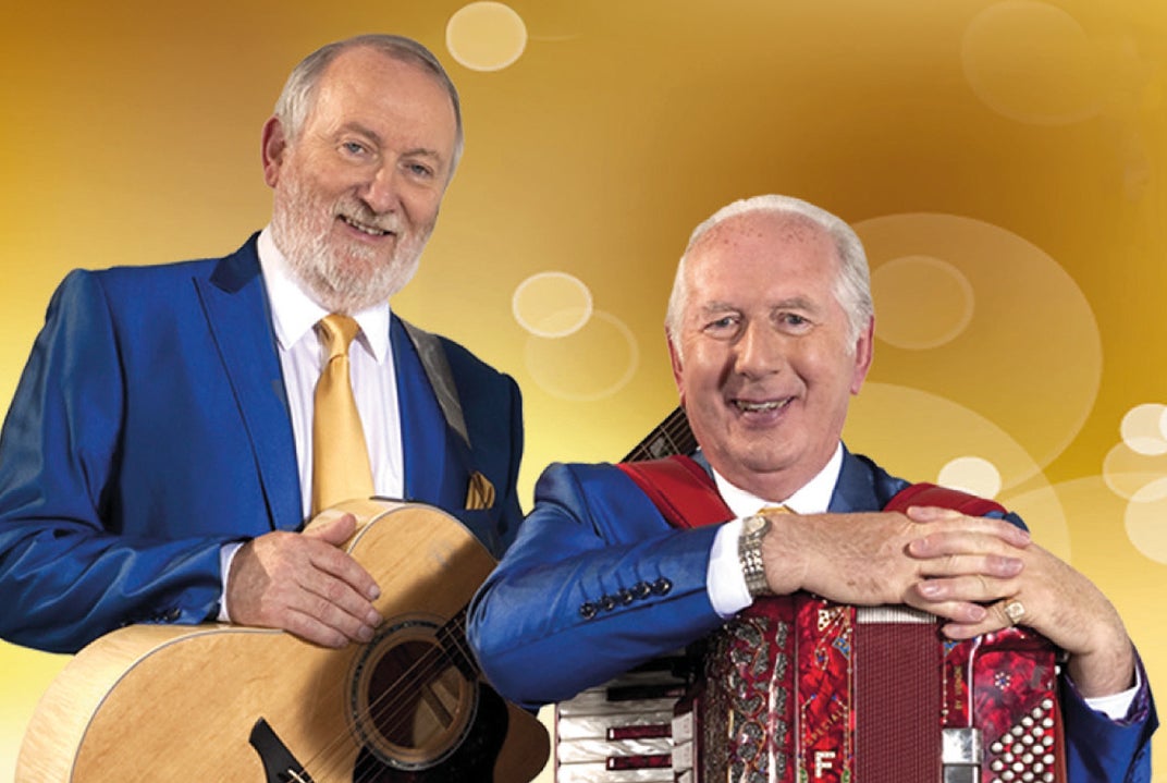 2 smiling older men resting their hands on musical instruments around their necks on straps