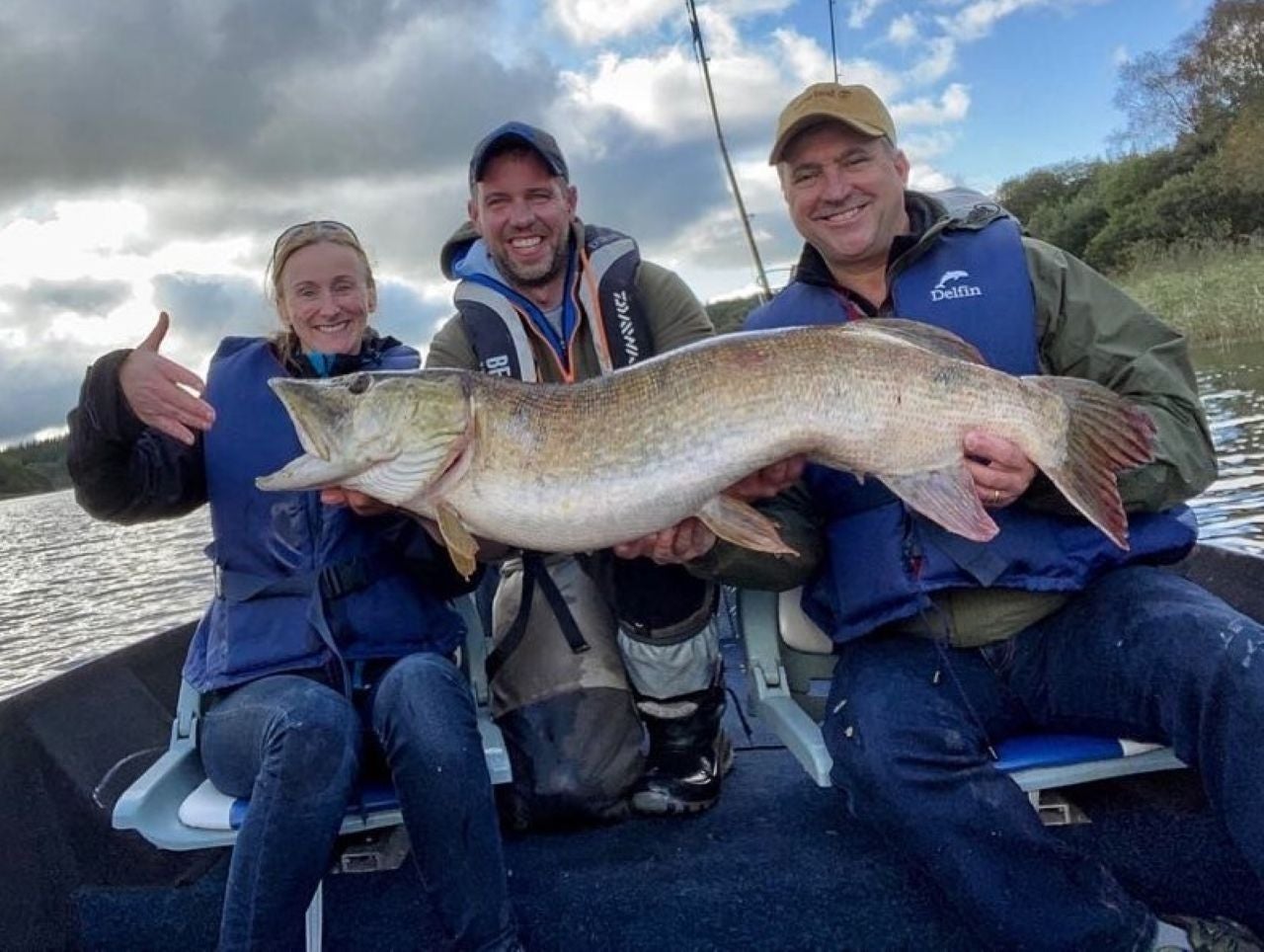 Three people in blue life vests sitting on a boat holding a big fish