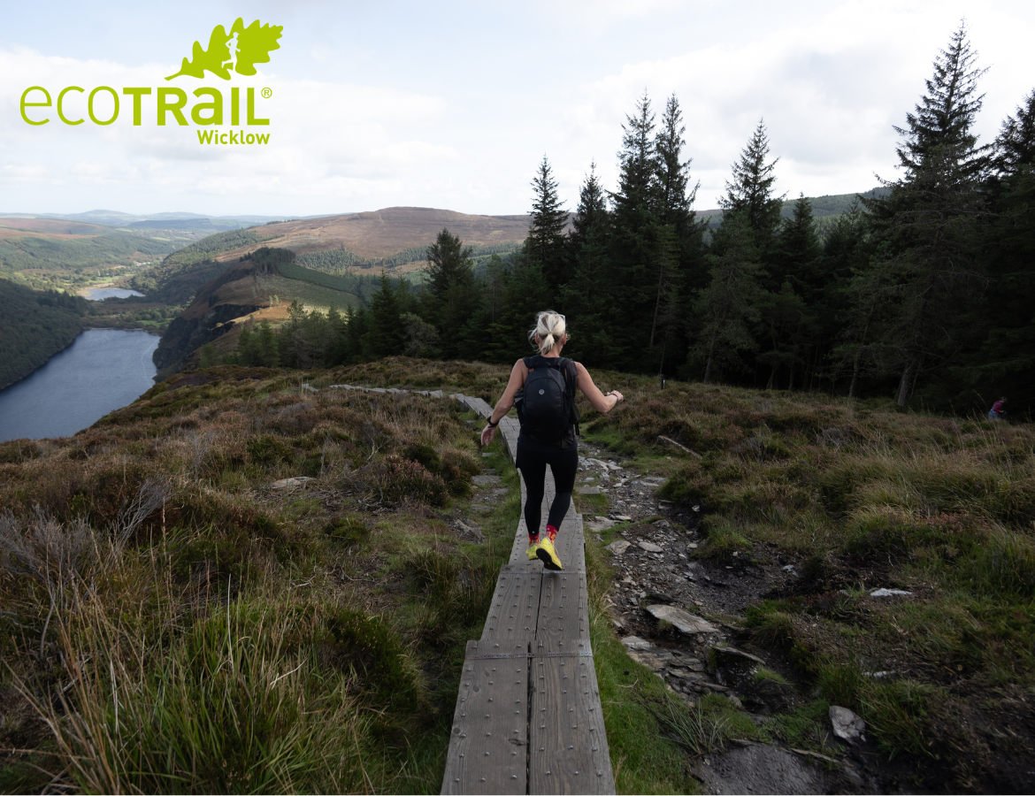EcoTrail Wicklow 2026 - a person running along a narrow wooden path on rough land with trees and hills in background.