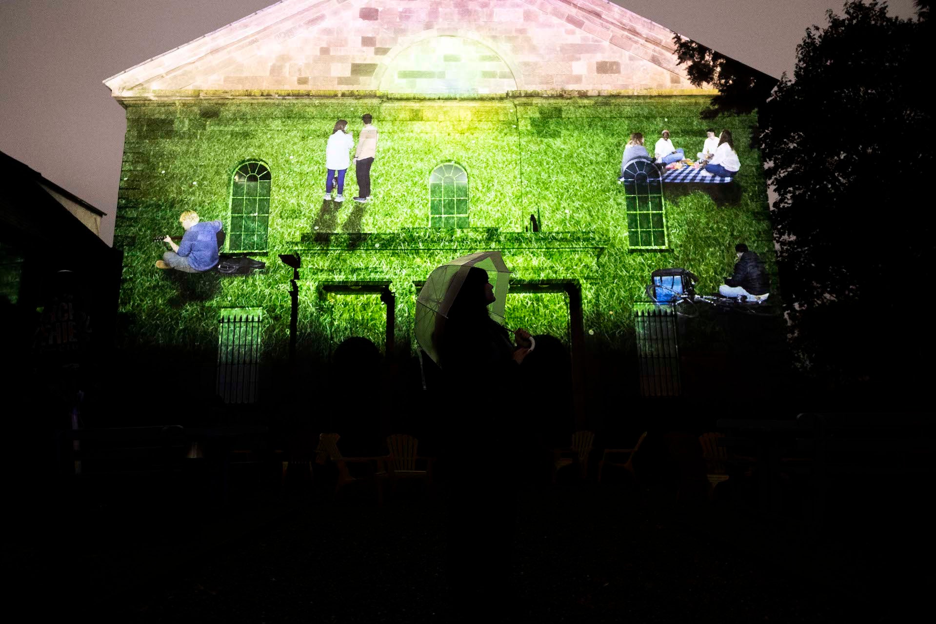 A projection of people on a field against the wall of a church. The silhouette of someone can be seen at the bottom of the image.