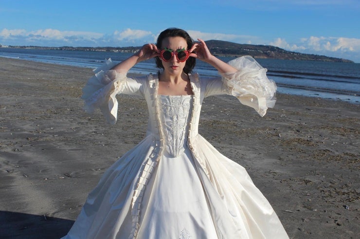 A woman in white, long 18th century dress holding on strawberry shaped sunglasses standing on a beach with the tide out.