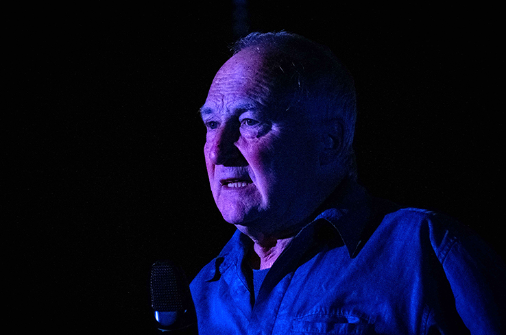 Actor Donal O'Kelly standing against a black background, face is lit by a purple-blue light