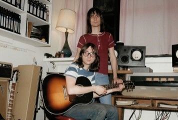 A standing and seated man with a guitar, beside a keyboard in a messy room