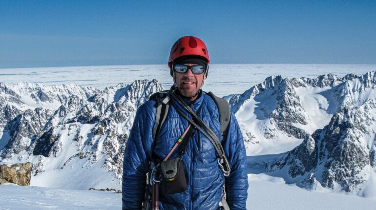 A man in outdoor clothing and mirrored sunglasses is standing against background of snowy mountains.