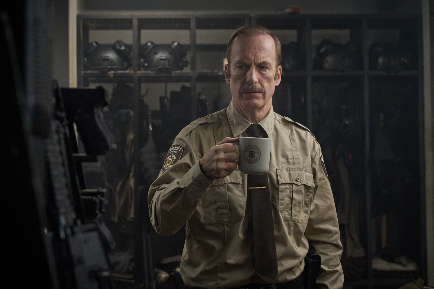 A man wearing a sheriff's uniform is in an armoury. He is holding a mug.