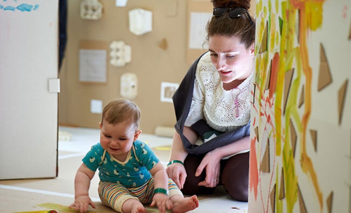 A smiling young baby seated on the floor looking down at something in front of them with an adult seated behind them.