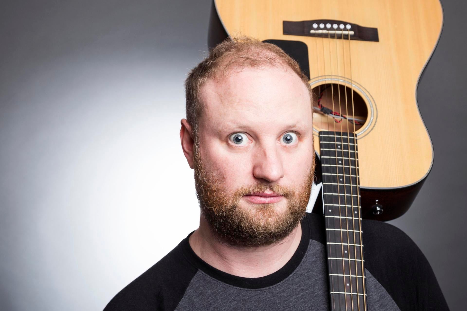 A wide eyed man is holding a guitar upside down resting on his left shoulder