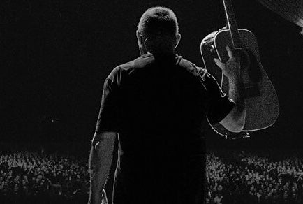 Darkly lit rear view of man holding up a guitar looking out over a large audience.