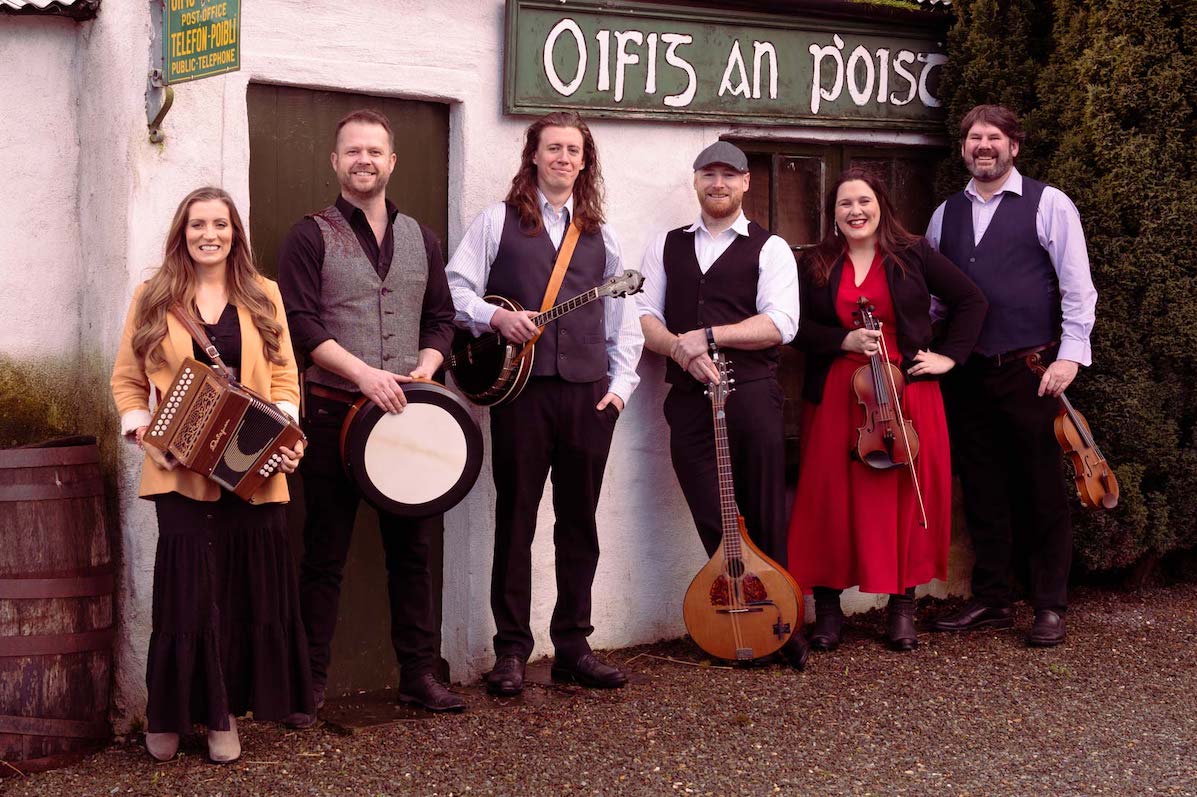 A line of 6 smiling men and women holding musical instruments standing in front of an old cottage