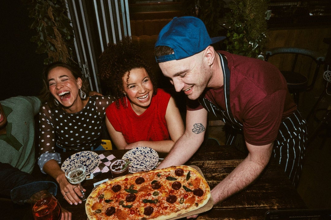 A waiter in an apron placing a large pizza on a table with smiling customers