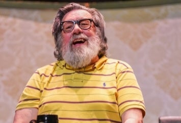 An Evening with Ricky Tomlinson, My Arse - a laughing older man with bushy beard seated with a mug on a table in front of him.