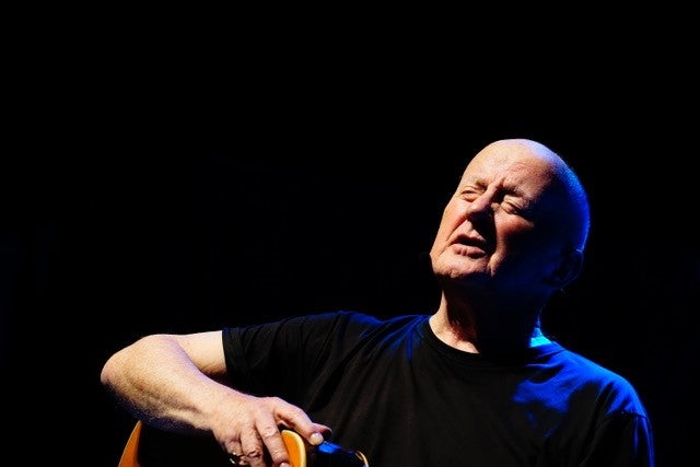 A bald man is singing with his eyes closed, a guitar on his lap.