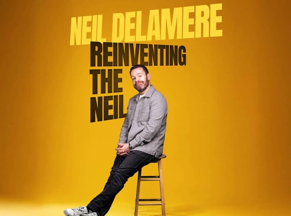 A smirking man perched on a tall stool against plain dull yellow background.