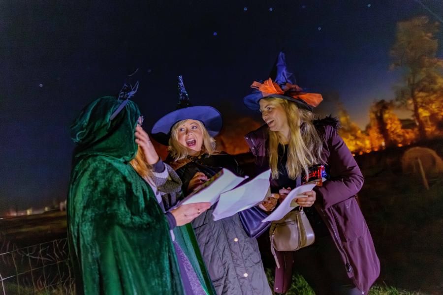 Adults in costume read their treasure hunt clues by torchlight at Nore Valley Park's Halloween Fire Trail, with warm lights glowing through the trees behind them.