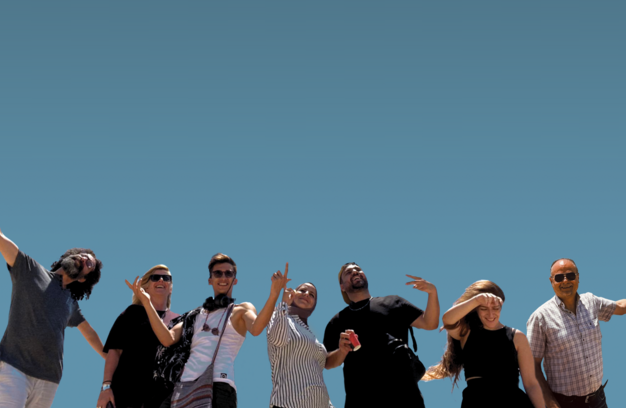 Looking up at a line of 7 happy people, some waving their arms, against a clear blue sky.