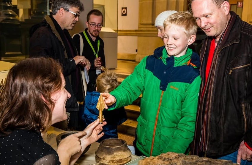 A adult and child touching the pointed end of an arrow head being help up by a woman seated at a table with other ancient artifacts.