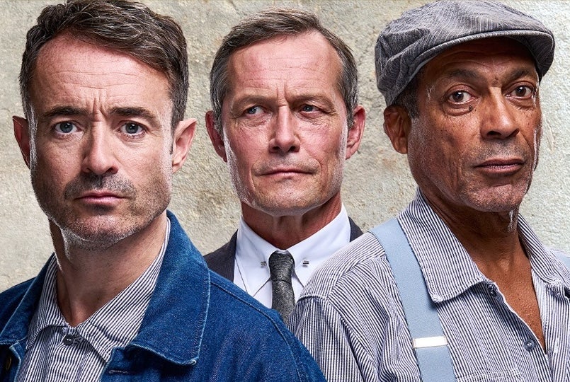 Headshots of 2 men in denim and stripy old shirts with man behind in formal shirt, tie and suit.