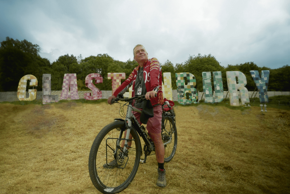A man looking wistful on a bike with large letters in the background spelling out a name of a town