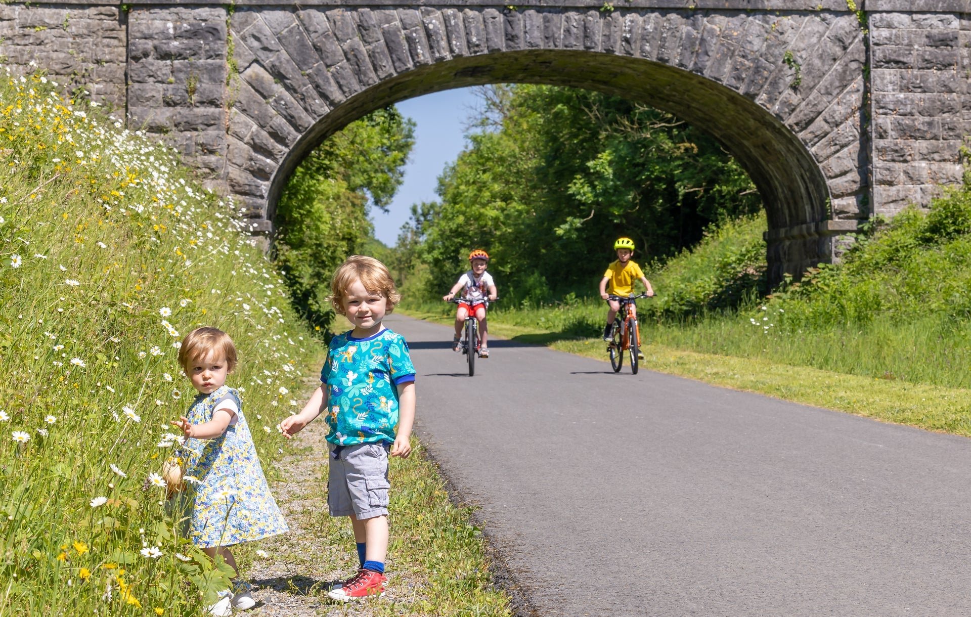 Two kids on bikes along the Old Rail Trail Greenway along with two kids that are picking flowers on the side of the trail